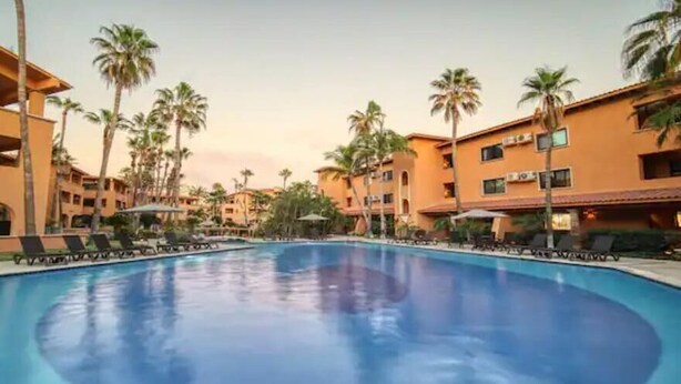 Zona de piscina de hotel con varios niveles de edificios, palmeras y sillas de descanso.