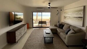Una sala de estar moderna con un televisor grande, un sofá, una mesa de centro y vistas a la playa a través de puertas de cristal.