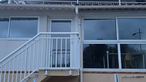 Un immeuble résidentiel à l’extérieur blanc, doté d’un balcon avec une balustrade blanche et d’une fenêtre donnant sur une pièce où l’on aperçoit une chaise et une plante.