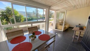 Un coin repas avec une table, des chaises et des sets de table rouges. On peut voir, à travers la fenêtre, de la verdure et un bâtiment.