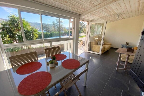 Un coin repas avec une table, des chaises et des sets de table rouges. On peut voir, à travers la fenêtre, de la verdure et un bâtiment.