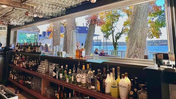 Un bar con vista a un lago, estantes con botellas de licores y copas colgantes.