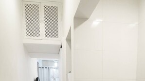 A narrow hallway with white tiled floors leading to a bathroom with a shower and toilet. A small trash bin is visible near the door. - Studio Sá Ferreira (Copacabana Rio de Janeiro)
