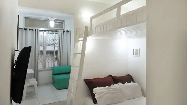 A room with a bed, a green chair, a small table, and a window with bars. - Studio Sá Ferreira (Copacabana Rio de Janeiro)