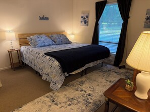 A bedroom with a bed, two bedside lamps, a small table with a lamp, a window with curtains, and artwork on the walls.