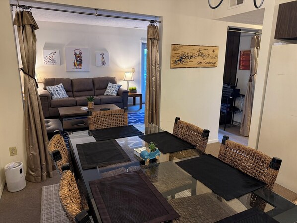 A living room with a sofa, coffee table, and chairs, a dining table with placemats, a lamp, and artwork on the walls.
