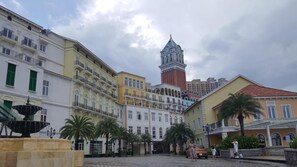 A historic square with a prominent clock tower, a fountain, and classic European architecture.