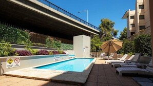 A swimming pool with lounge chairs and umbrellas, surrounded by greenery and a building. - Urban CBD Perth A GEM 1Bedroom Apartment (Perth)