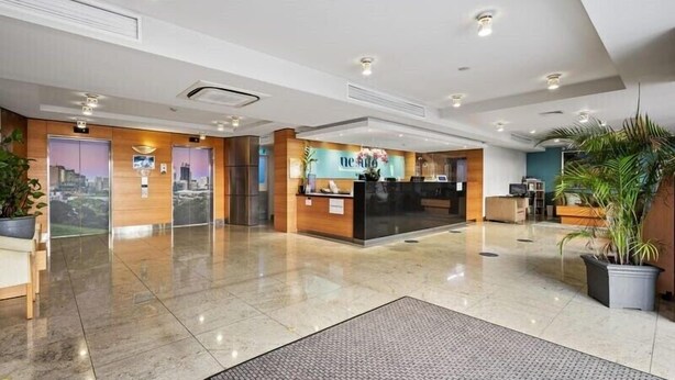 A modern hotel lobby with a reception desk, a large potted plant, and a cityscape view through a glass door. - Urban CBD Perth A GEM 1Bedroom Apartment (Perth)
