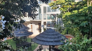 An outdoor patio area with thatched umbrellas, a glass table, and chairs, surrounded by lush greenery and a building in the background. - Coastal Salty Kiss Sleeps 4! (Scarborough)