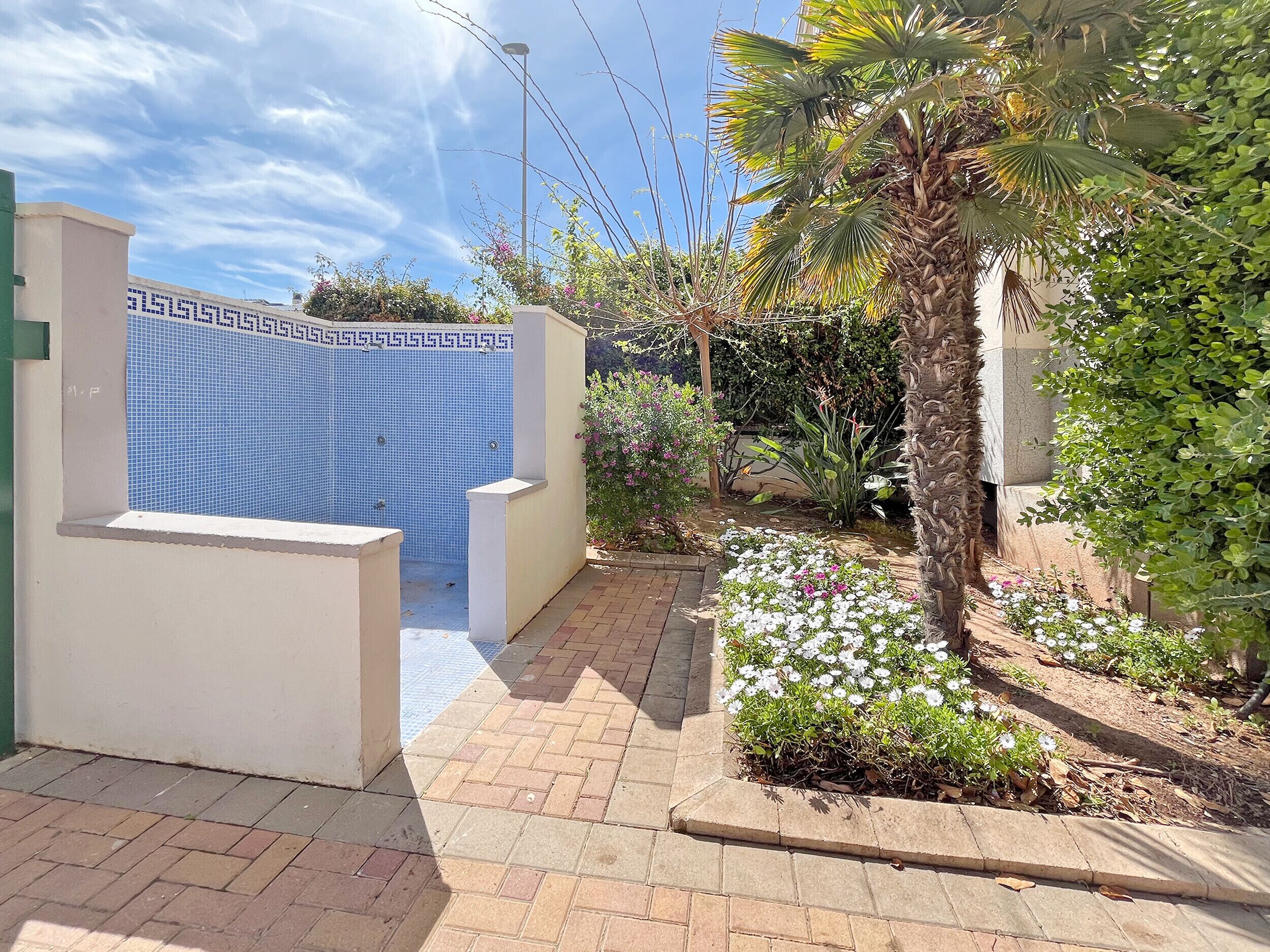 Una entrada con reja, pared de azulejos, camino empedrado y jardín con flores y plantas.