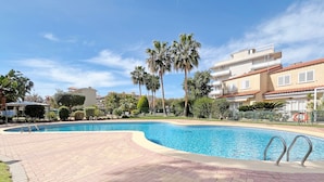 A swimming pool with a brick deck, surrounded by palm trees and a building with a red-tiled roof. - Global Vacacional: Practical flat in Canet Beach (Canet d´en berenguer)