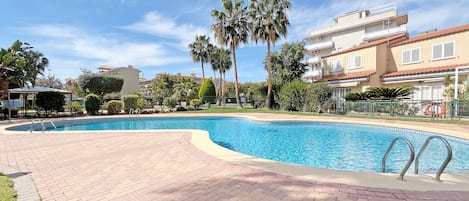 Una piscina con borde de ladrillo, rodeada de palmeras y un edificio con tejado rojo.