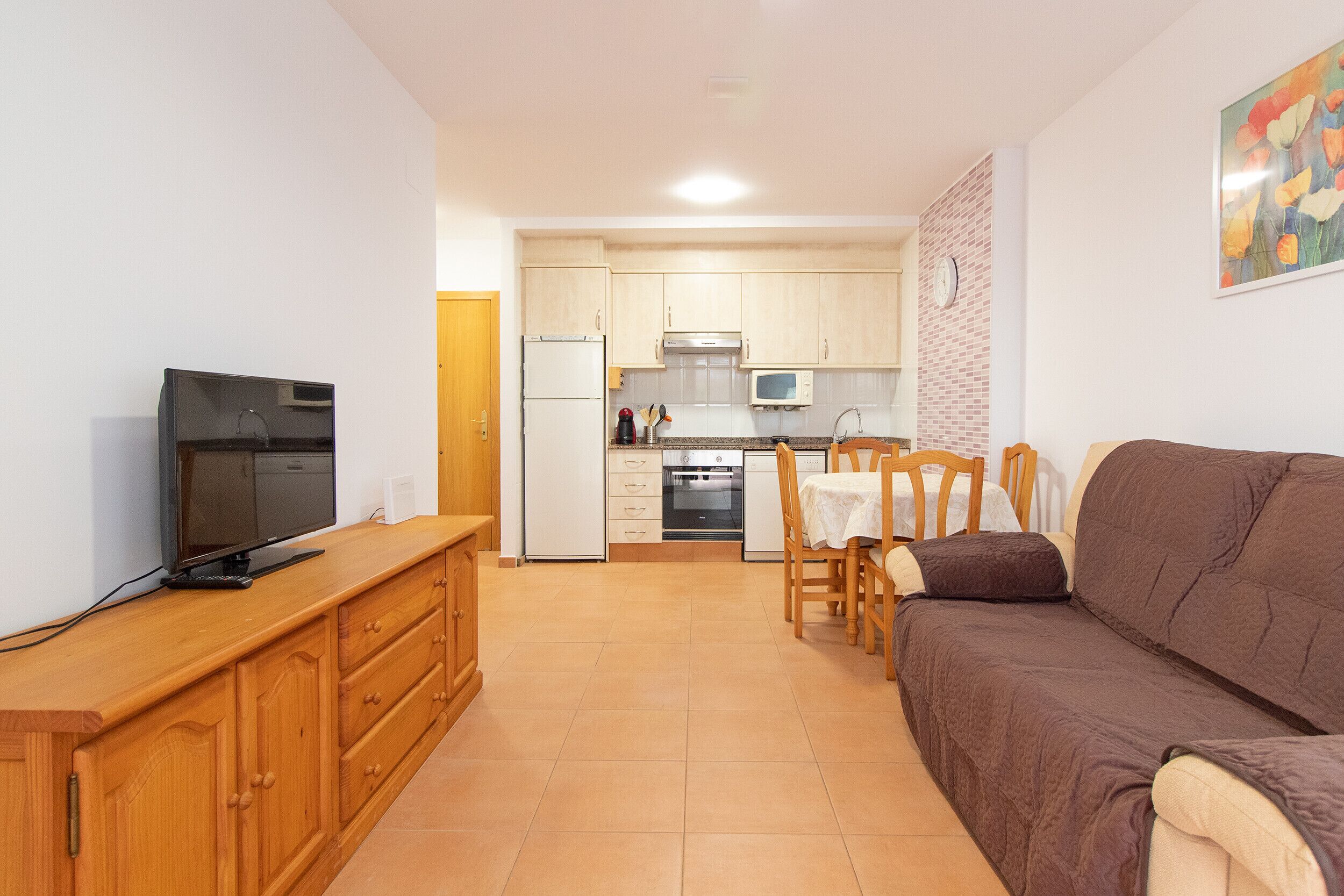 A compact living space with a kitchenette, a television on a wooden cabinet, a brown sofa, and a dining table with chairs.