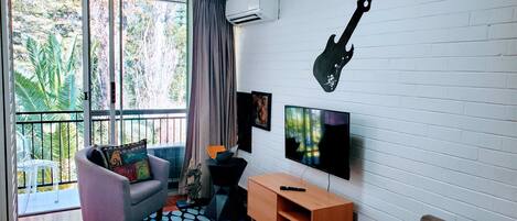 A modern living room with a grey sofa, a mounted guitar, a TV, and a balcony with a view of greenery.