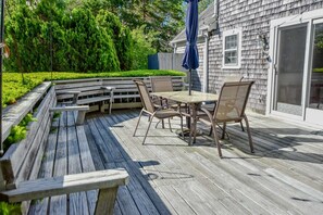 A wooden deck with a glass-top table and chairs, surrounded by a wooden bench and a small tree. - Walk to Skaket Beach, Great Backyard! (Orleans)