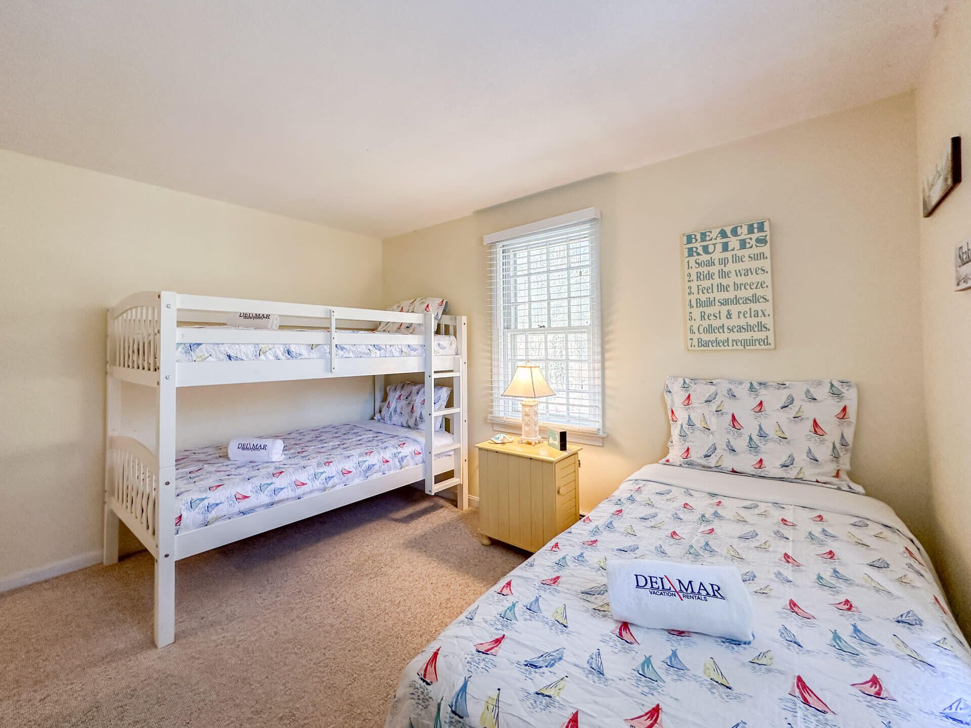 A bedroom with bunk beds, a single bed with a patterned duvet, a bedside lamp, a window with blinds, and a wall sign with a list of items.