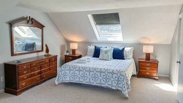 A bedroom with a sloped ceiling, a skylight, a bed with patterned bedding, two bedside tables with lamps, and a dresser.
