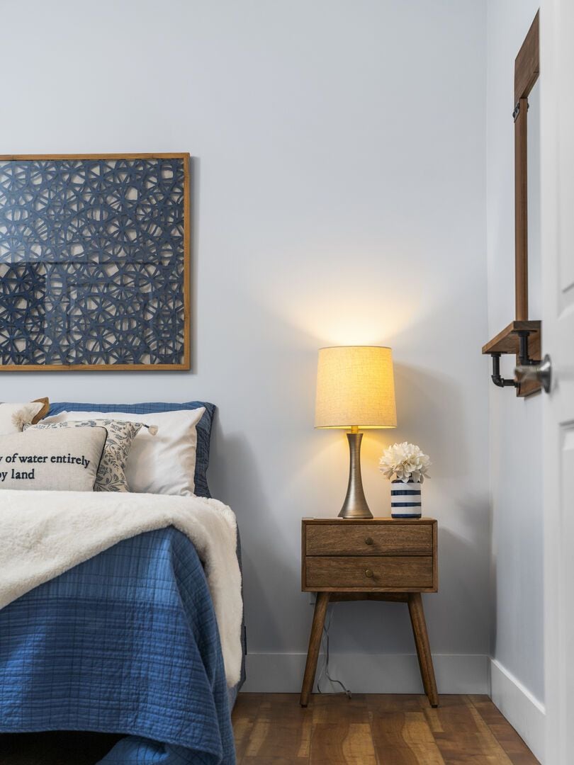 A bedroom with a bed, a bedside table with a lamp, a decorative wall piece, and a door.