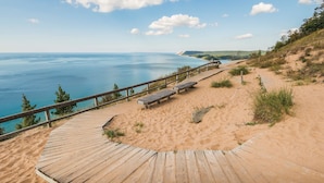 A wooden boardwalk leading to a sandy beach with benches, overlooking a calm sea and distant hills. - The Heart of TC 2BDR Condo Unit 115 (Traverse City)
