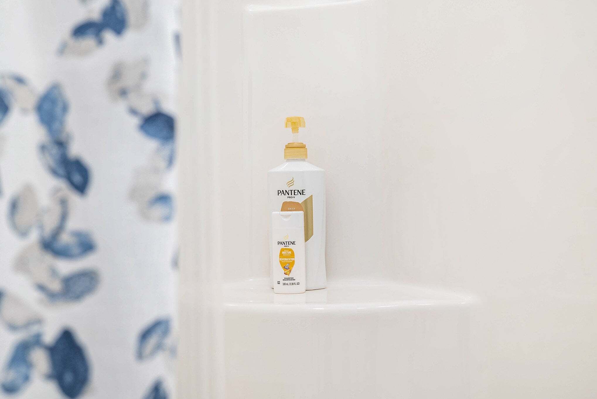 A bathroom with a white shower curtain and a Pantene shampoo bottle on a white shelf.