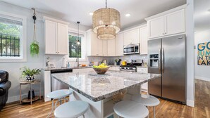 A modern kitchen with white cabinets, a stainless steel refrigerator, a hanging light fixture, and a central island with bar stools. - The Heart of TC 2BDR Condo Unit 115 (Traverse City)