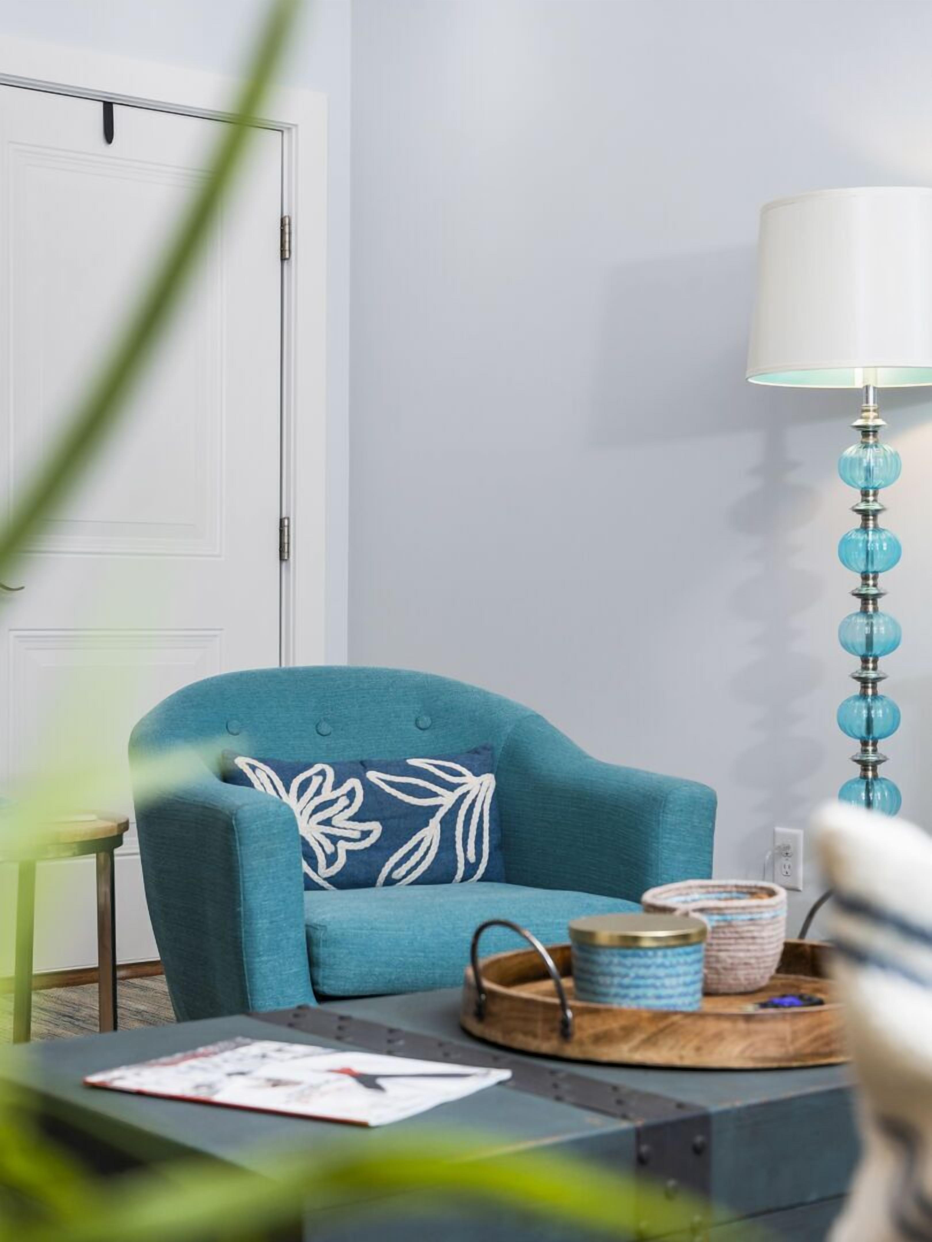 Een kamer met een blauwgroene fauteuil, een witte deur, een lamp met blauw glas en een klein tafeltje met een dienblad.