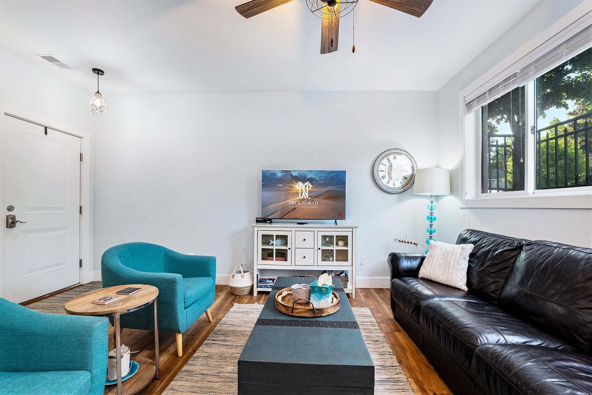 A living room with a black leather couch, teal armchairs, a coffee table, a TV, and a window with a view.