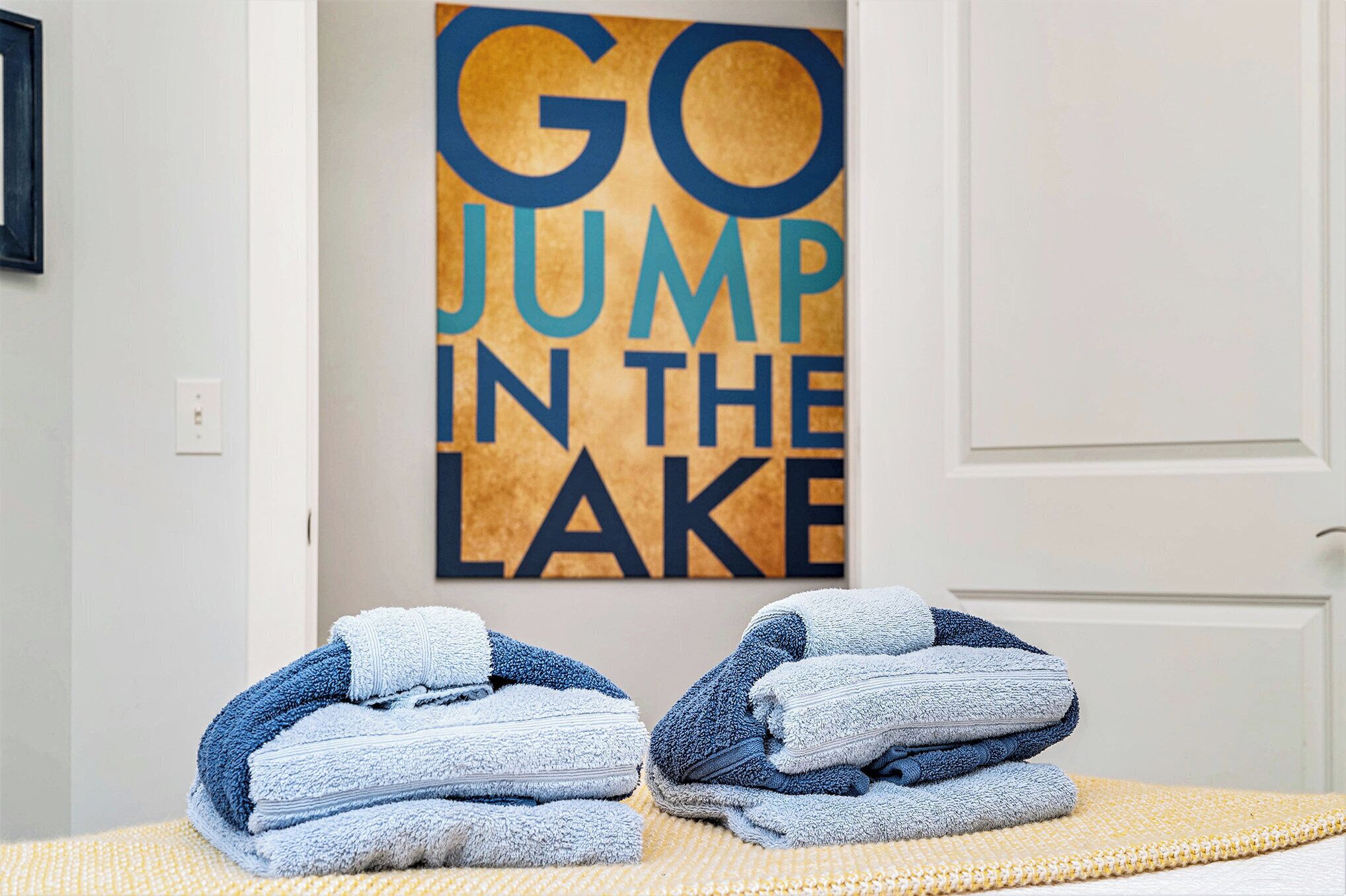 A bathroom with towels stacked on a yellow mat, a sign that reads "GO JUMP IN THE LAKE", and a white door.