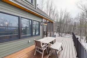 Eine Holzterrasse mit Tisch und Stühlen, großen Fenstern und Blick auf Bäume.