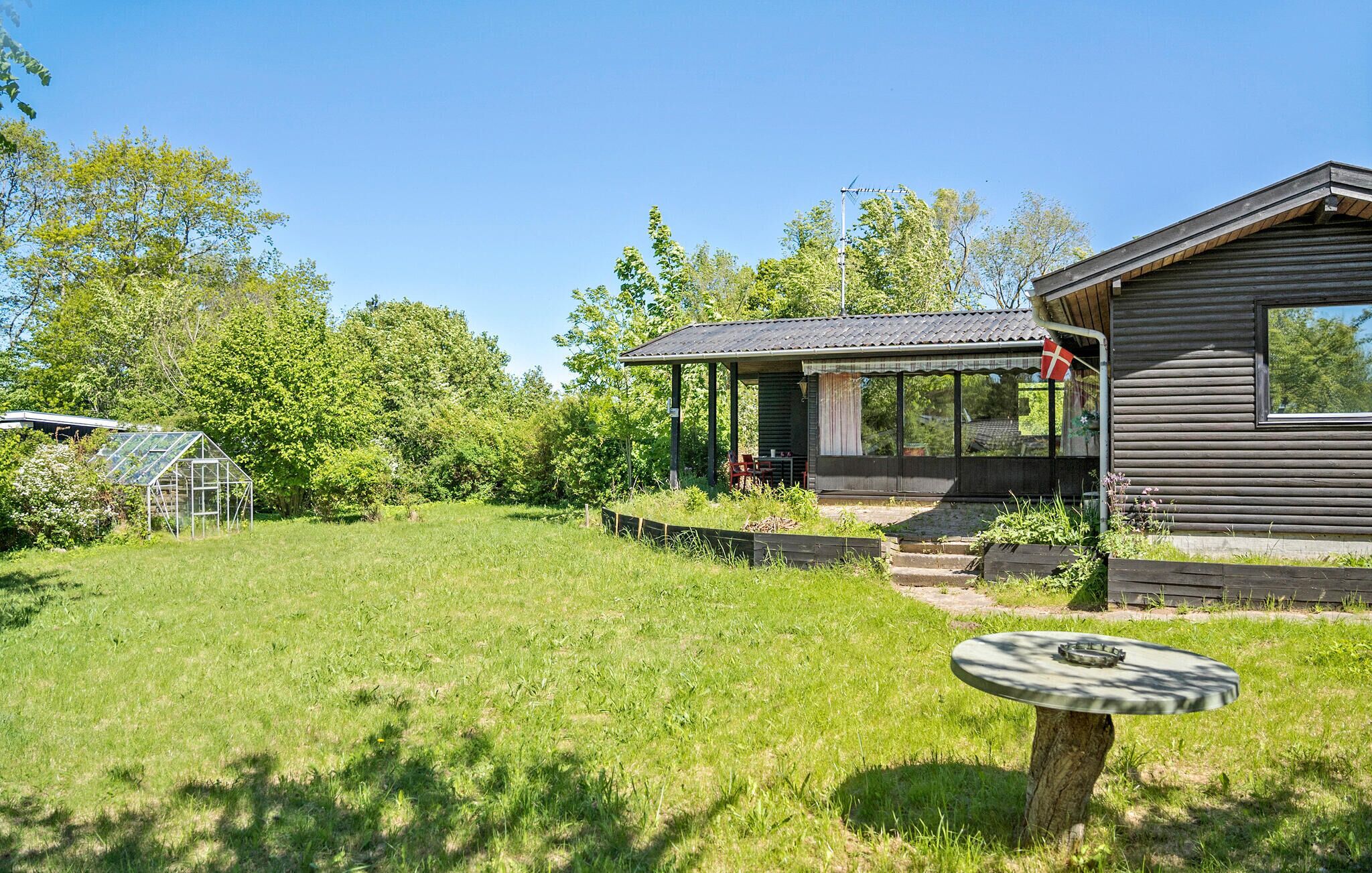 Een houten hut met een overdekte veranda en een kas, gelegen in een weelderige groene omgeving.