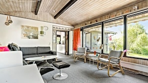 Un salon moderne avec un canapé, une table basse et des fauteuils, de grandes fenêtres donnant sur un paysage verdoyant et un plafond en bois.