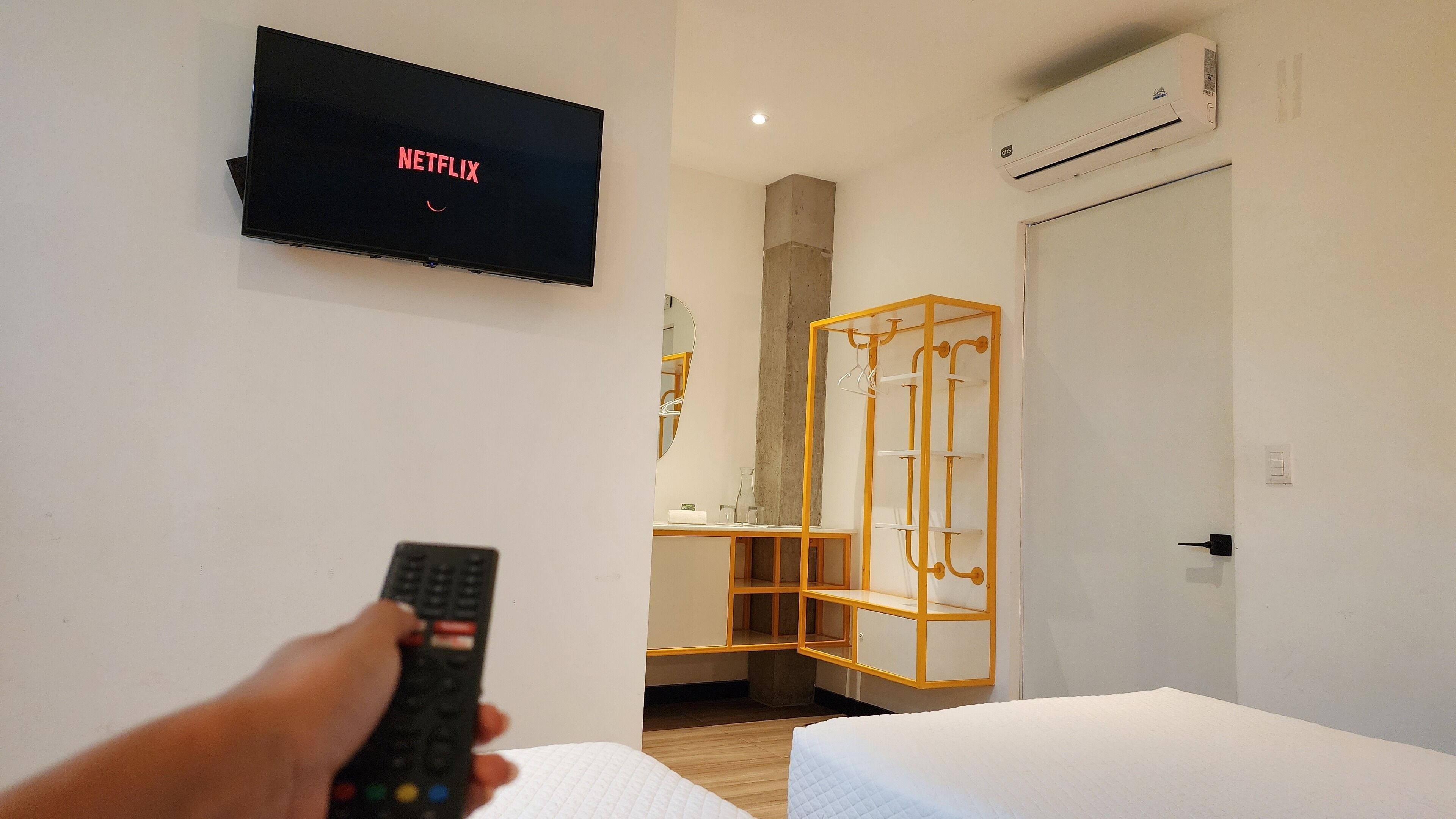 Una habitación de hotel con una cama, un televisor con el logotipo de Netflix y un armario con espejo, además de un equipo de aire acondicionado.