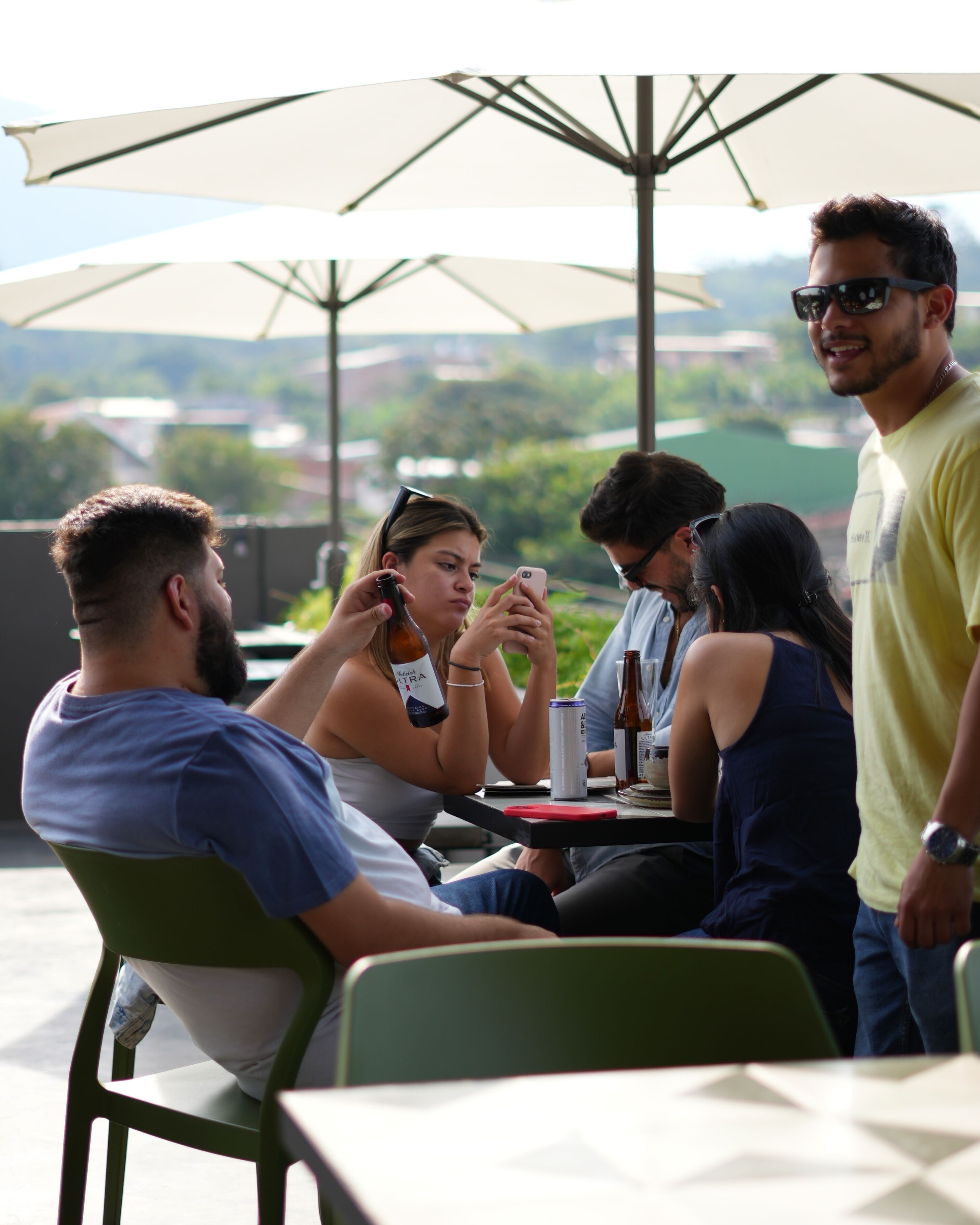 A group of people sitting at an outdoor table with umbrellas, engaged in conversation and using their phones.