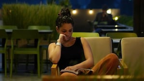 Iemand zit op een stoel bij een café met terras, met een boek, omringd door groen en tafels.