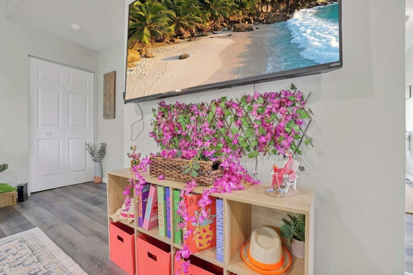 A living room with a TV displaying a beach scene, a bookshelf with books and decorative items, and a wall-mounted plant.