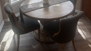 En spiseplads med et rundt bord, seks stole og en potteplante. Tre vinduer giver naturligt lys og udsigt til det fri.