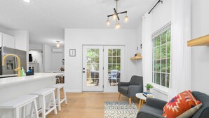 A modern kitchen with white cabinets, a bar area with white stools, a grey sofa with a colorful cushion, and a patterned rug. - Gorgeous 2BDR Home near Downtown (Traverse City)