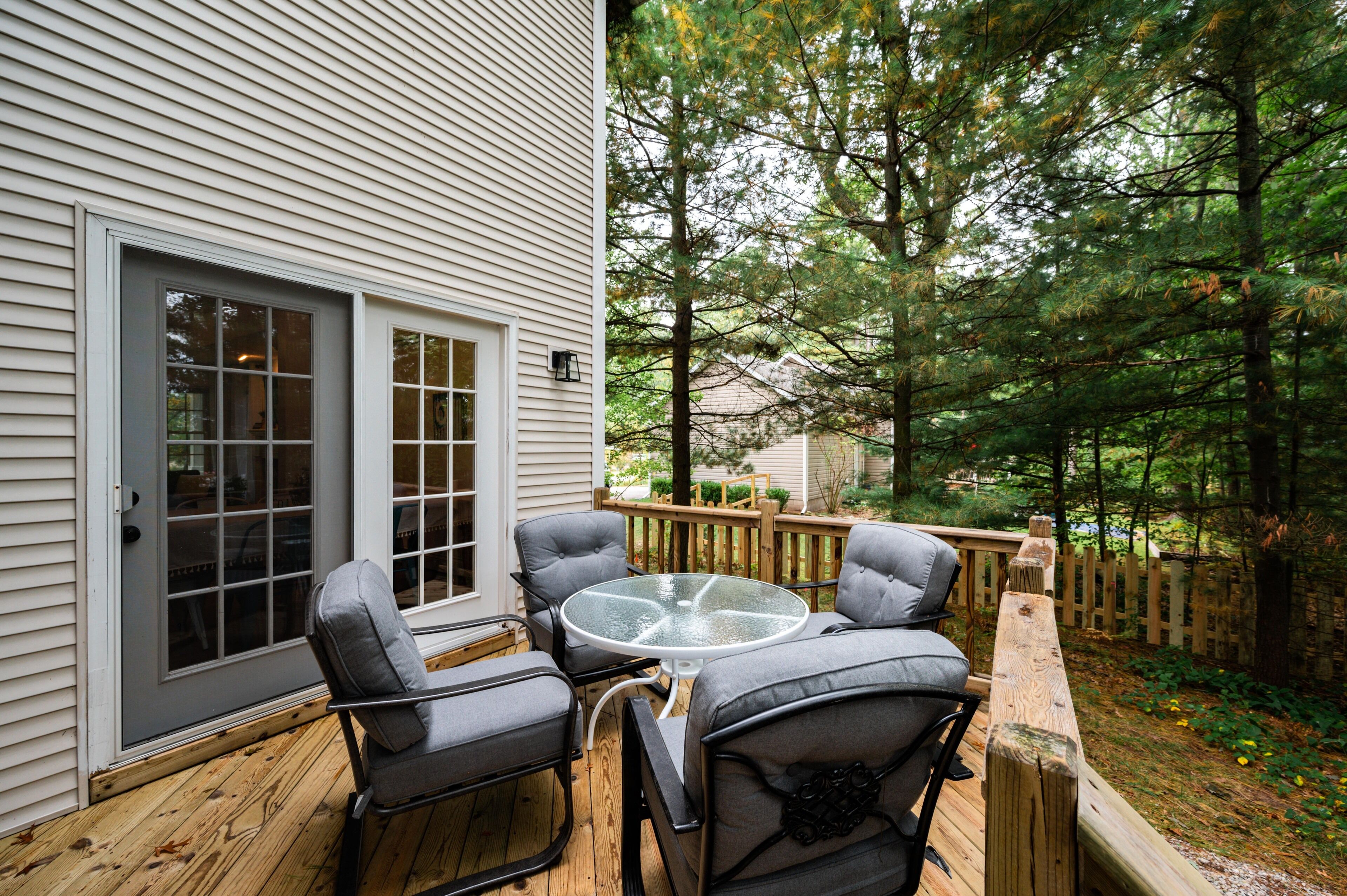 A deck with four gray chairs and a glass-top table, surrounded by a wooden deck and a wooden fence.