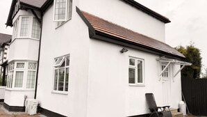 A white two-story house with a tiled roof, a bay window, and a small outdoor seating area with a chair and a table. - Nice and Cosy one bed flat
 (Carshalton)