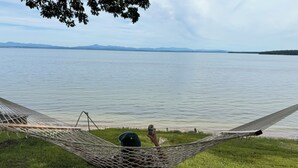 A hammock on a grassy area overlooking a calm lake with distant mountains. - A New York camp on Lake Champlain, private pebble beach frontage. (Keeseville)