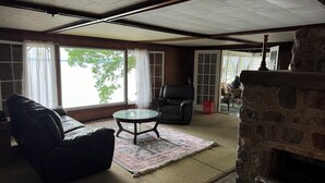 A living room with a fireplace, a leather sofa, a glass coffee table, and a view of the outdoors through large windows. - A New York camp on Lake Champlain, private pebble beach frontage. (Keeseville)
