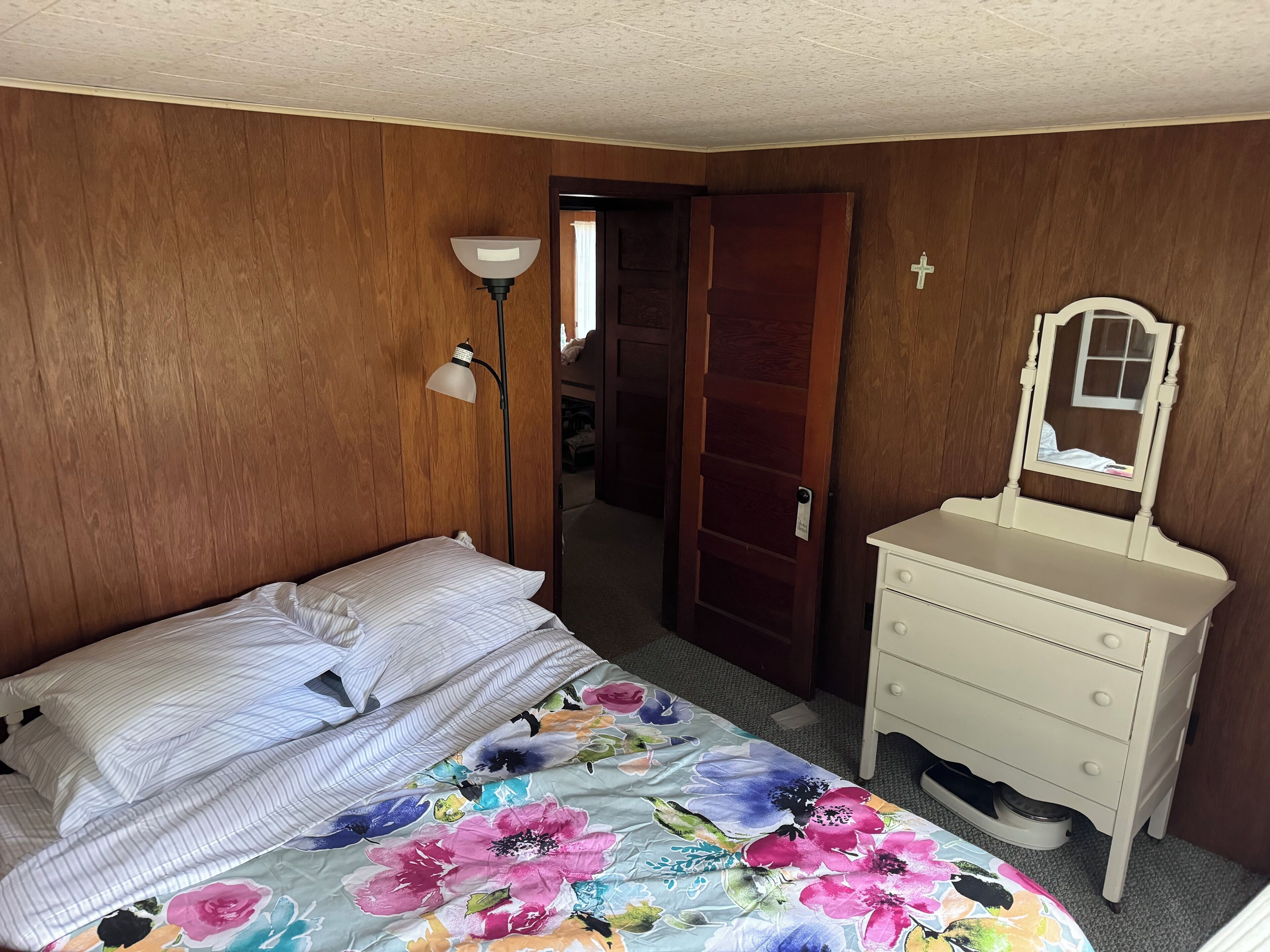 Una camera da letto con un letto, un comodino, una cassettiera, uno specchio e una porta che dà su un'altra stanza.