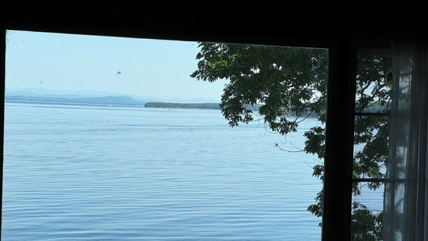A view of a calm lake with distant mountains and a tree in the foreground. - A New York camp on Lake Champlain, private pebble beach frontage. (Keeseville)