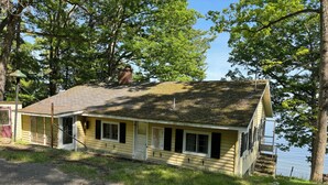 A small, single-story cabin with a porch, a chimney, and a view of a lake. - A New York camp on Lake Champlain, private pebble beach frontage. (Keeseville)