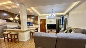 Una sala moderna con un sofá gris, suelo de madera y una zona de cocina con armarios oscuros e iluminación colgante.