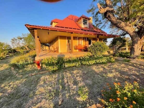Stroll through the serene garden, surrounded by lush greenery and a majestic tree, to the charming single-story house with a covered porch.