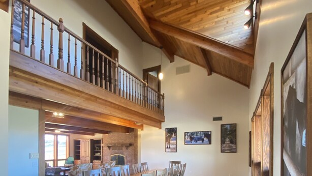 A spacious dining area with a high ceiling and wooden beams, a balcony with a railing, and several framed pictures on the walls.