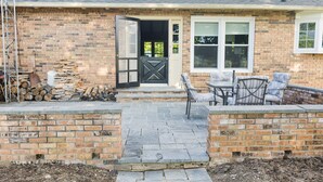 A brick house with a stone patio, a black door, and a small outdoor seating area with chairs.