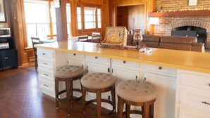A kitchen with a central island, wooden stools, a brick fireplace, and a dining area with chairs and a table.